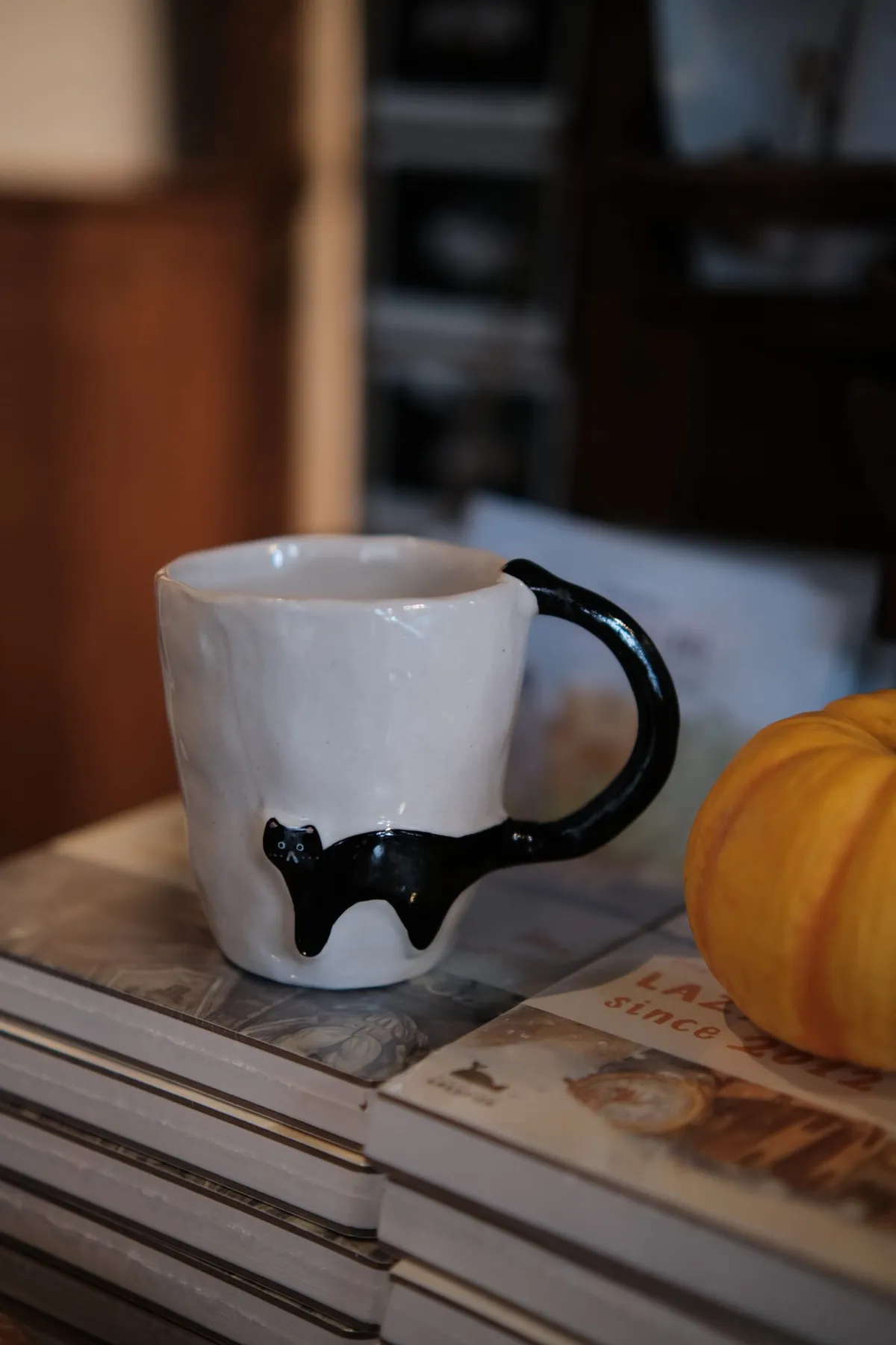 Black Cat mug on a stack of books beside a small pumpkin, the cat's tail curling up into the handle