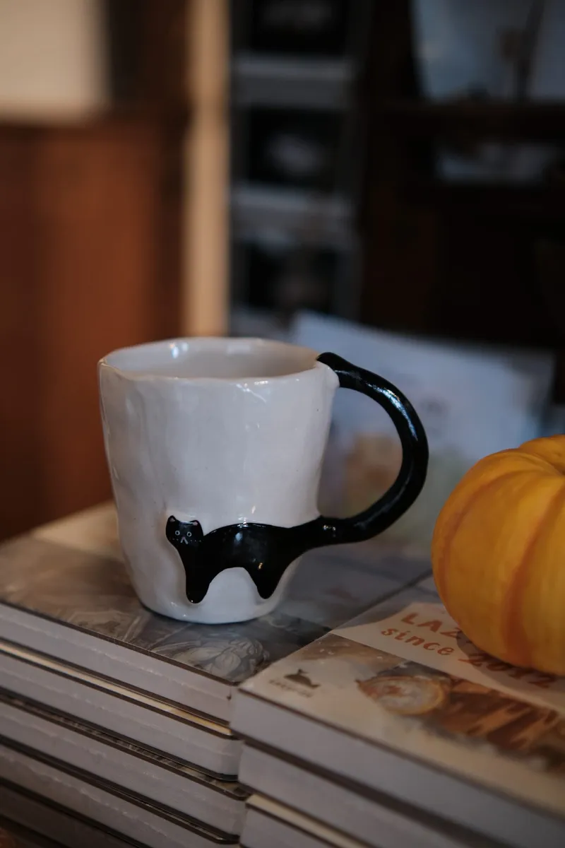 Black Cat mug on a stack of books beside a small pumpkin, the cat's tail curling up into the handle