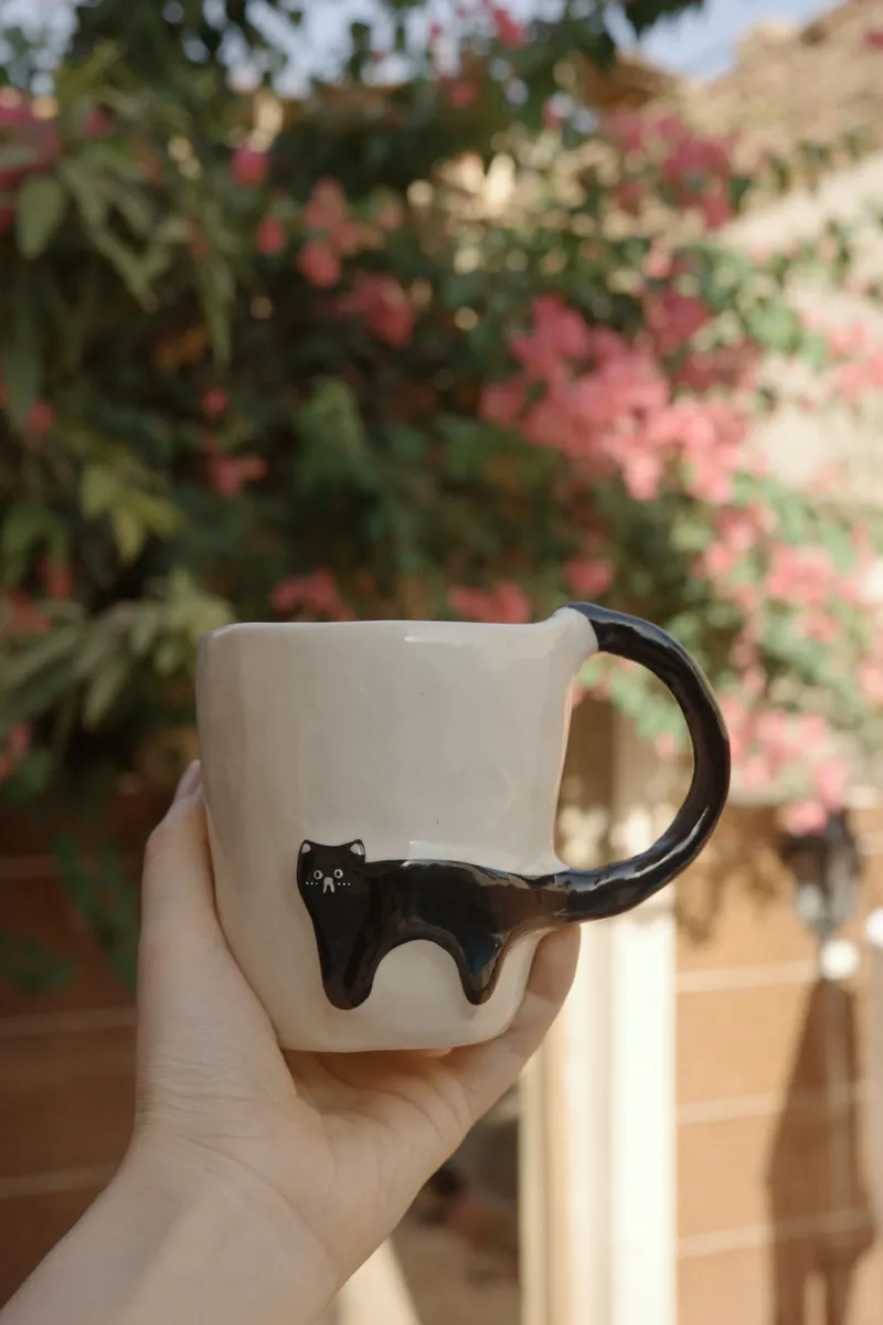 Black Cat mug held up in one hand in front of pink bougainvillea