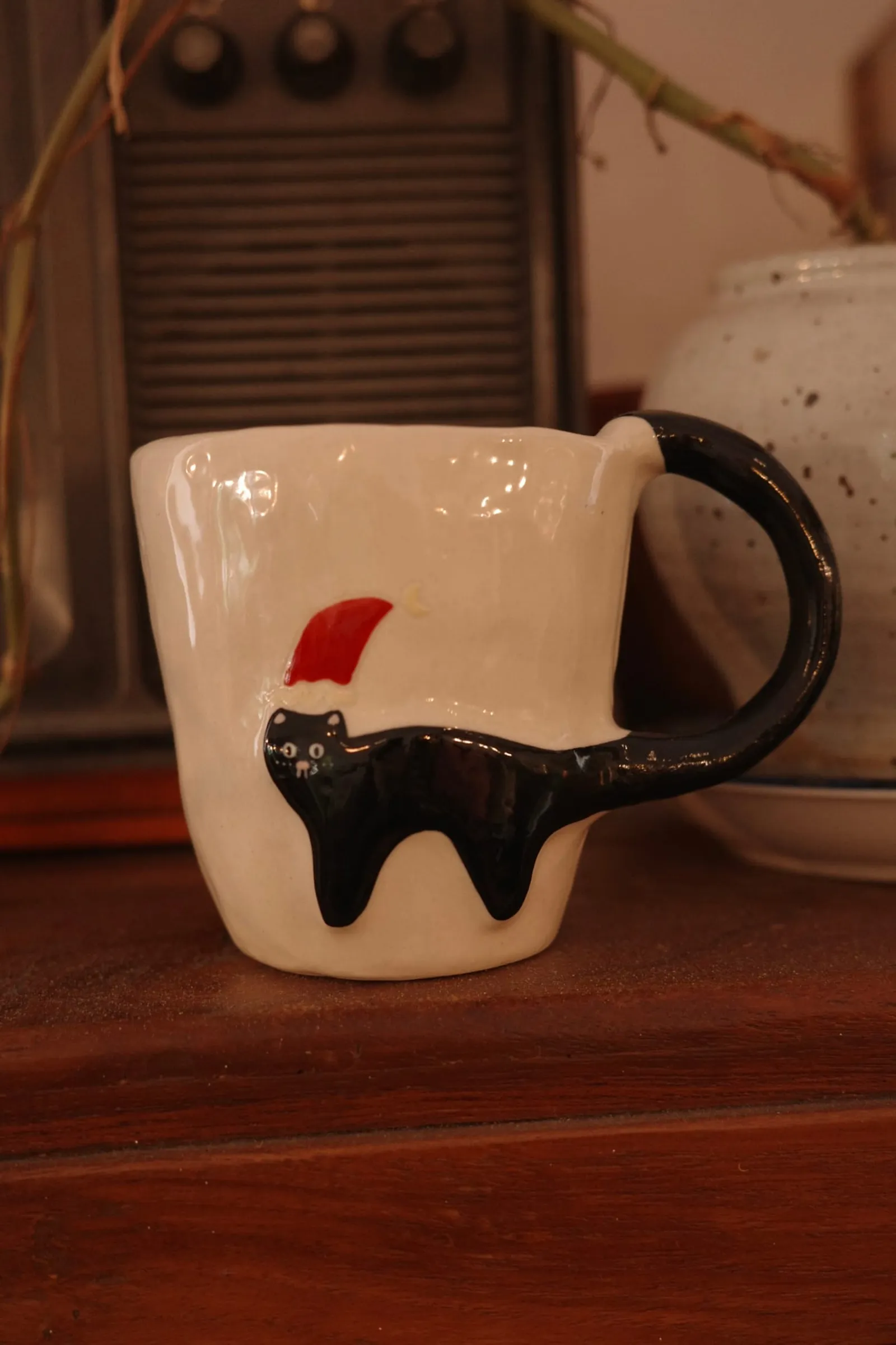 Black cat in a red Christmas hat on a white mug, on a dark wooden counter in warm light