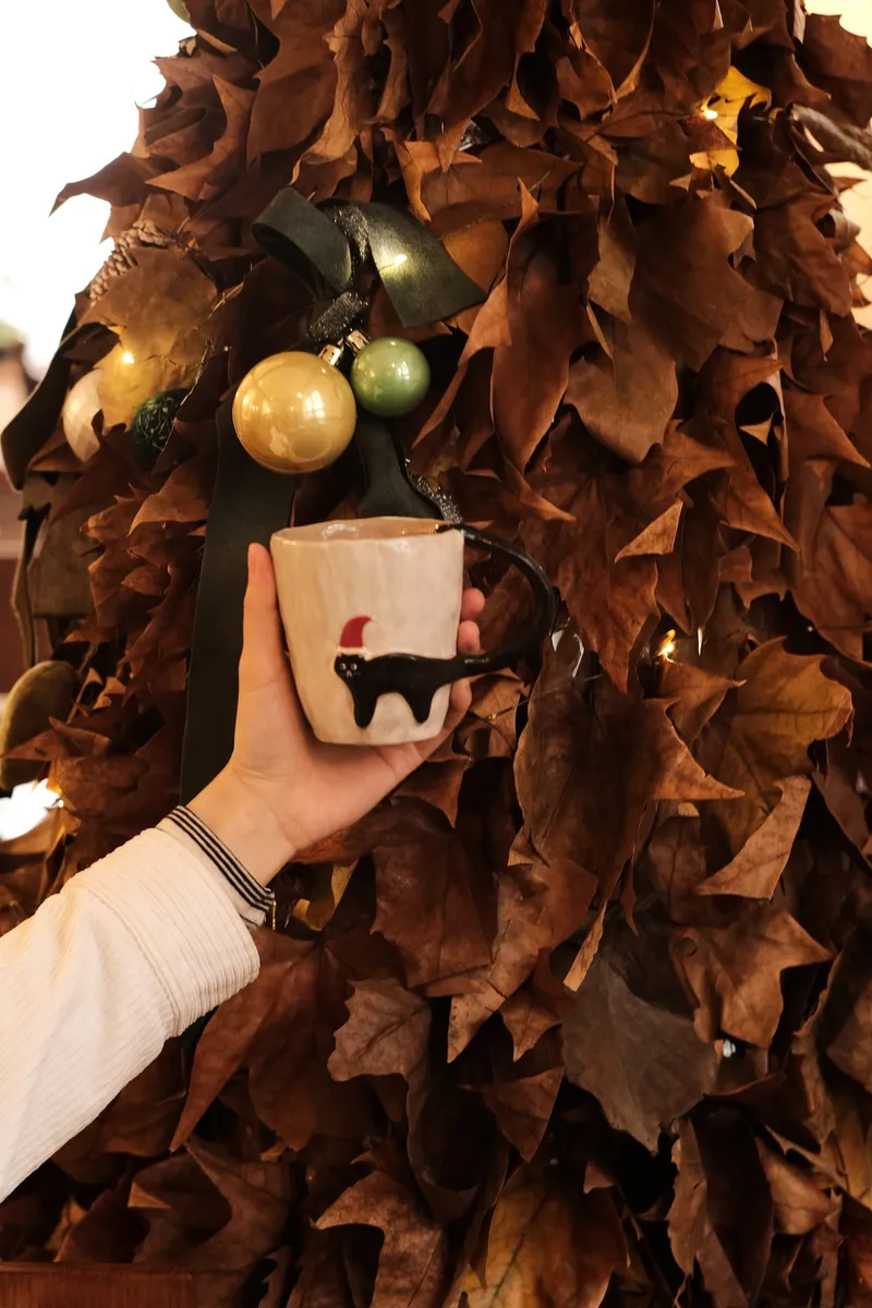 Christmas black cat mug held up in front of a dried-leaf Christmas tree