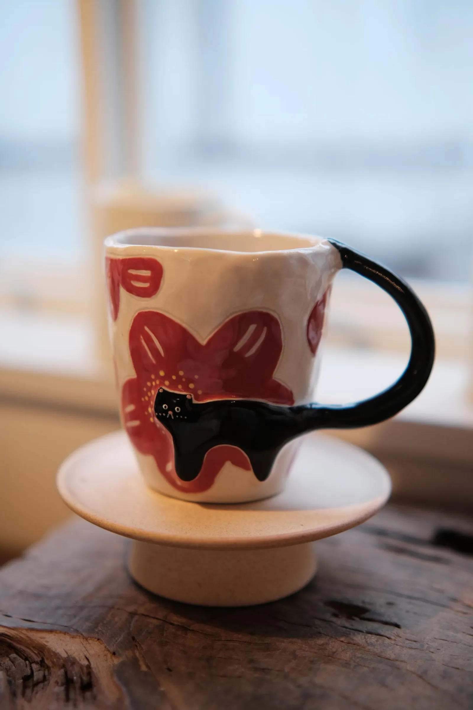 Black cat mug with red flowers on a ceramic stand in soft window light