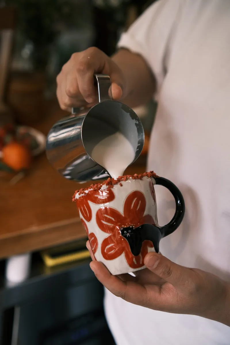 A barista pouring milk into the red flowers mug at a coffee bar