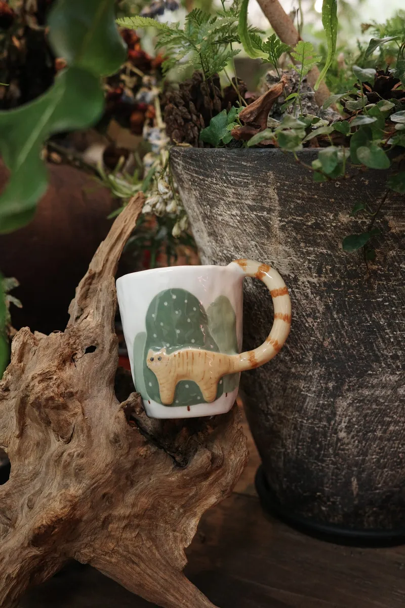 Orange cat mug with a green sky background sitting on driftwood among plants
